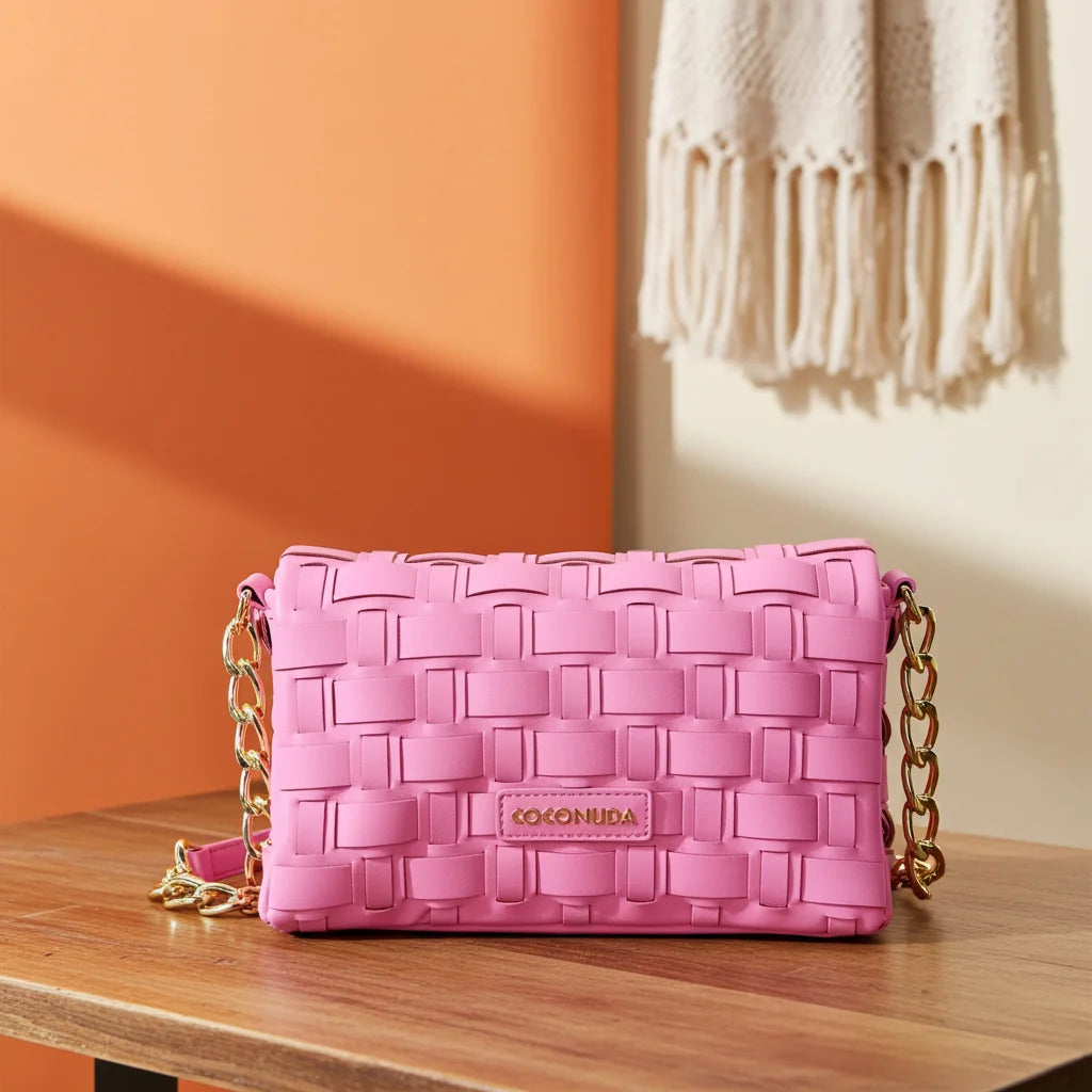 Pink woven handbag with gold chain on a wooden surface against an orange wall.